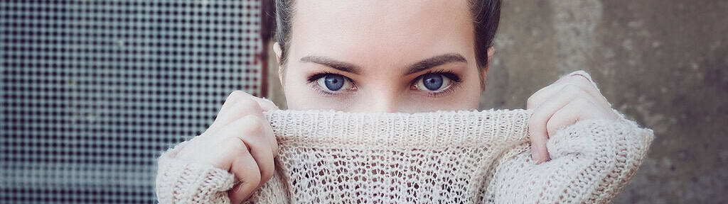 A woman with intense blue eyes, partially covering her face with a knitted sweater, symbolizing introspection and vulnerability, relevant to EMDR Therapy for Trauma and PTSD.