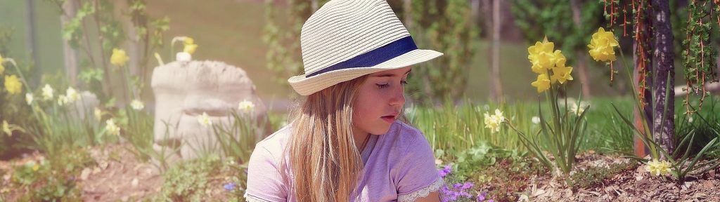 A young girl wearing a straw hat and a light purple shirt sits cross-legged on a small suitcase in a garden with colourful flowers. She appears to be deep in thought, symbolising the calm and introspective nature of EMDR therapy for children.