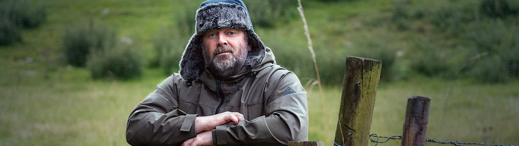 A man wearing a winter hat and jacket leaning on a wooden fence in a rural setting, symbolizing contemplation and decision-making, possibly reflecting on in-person vs online EMDR therapy options.