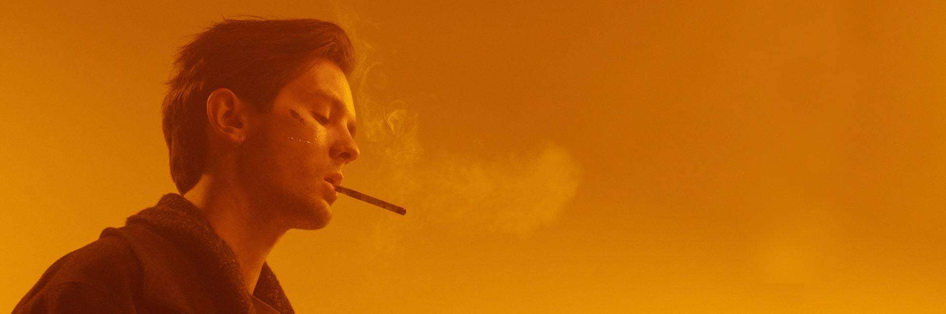 A man with a pensive expression, exhales smoke against an orange-hued background, relating to AERCS addiction counselling.