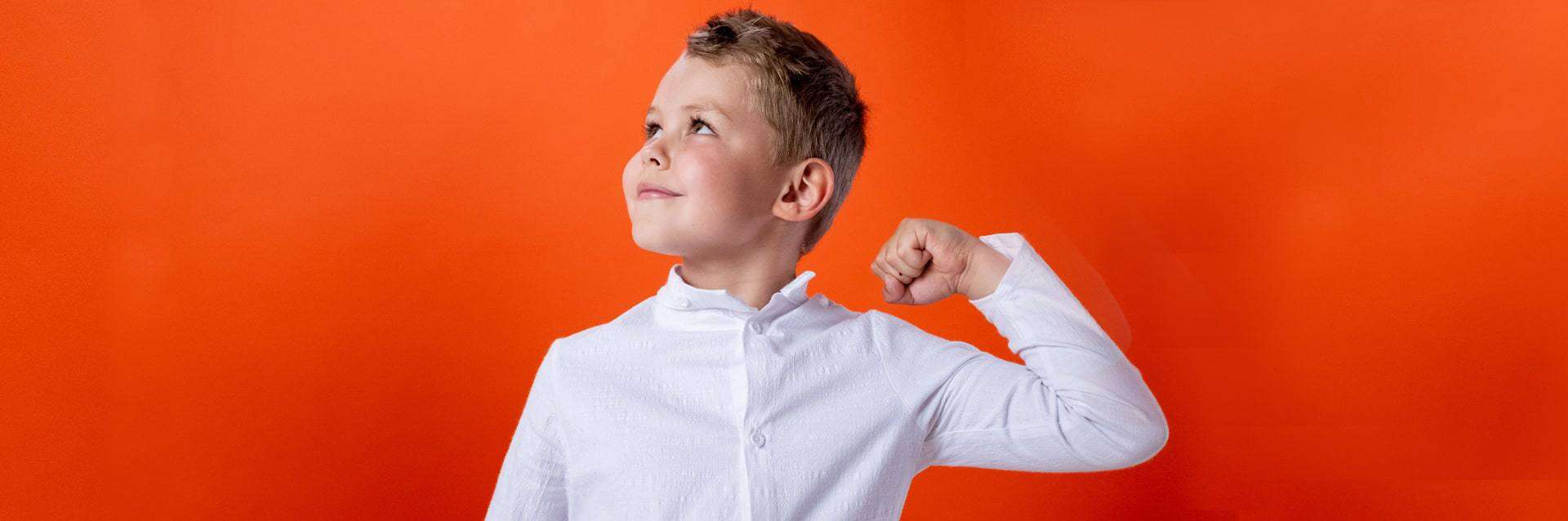 Confident young boy striking a strong pose against a bright orange background, symbolizing the self-esteem and emotional growth fostered through play therapy. Confident young boy striking a strong pose against a bright orange background, symbolizing the self-esteem and emotional growth fostered through play therapy.