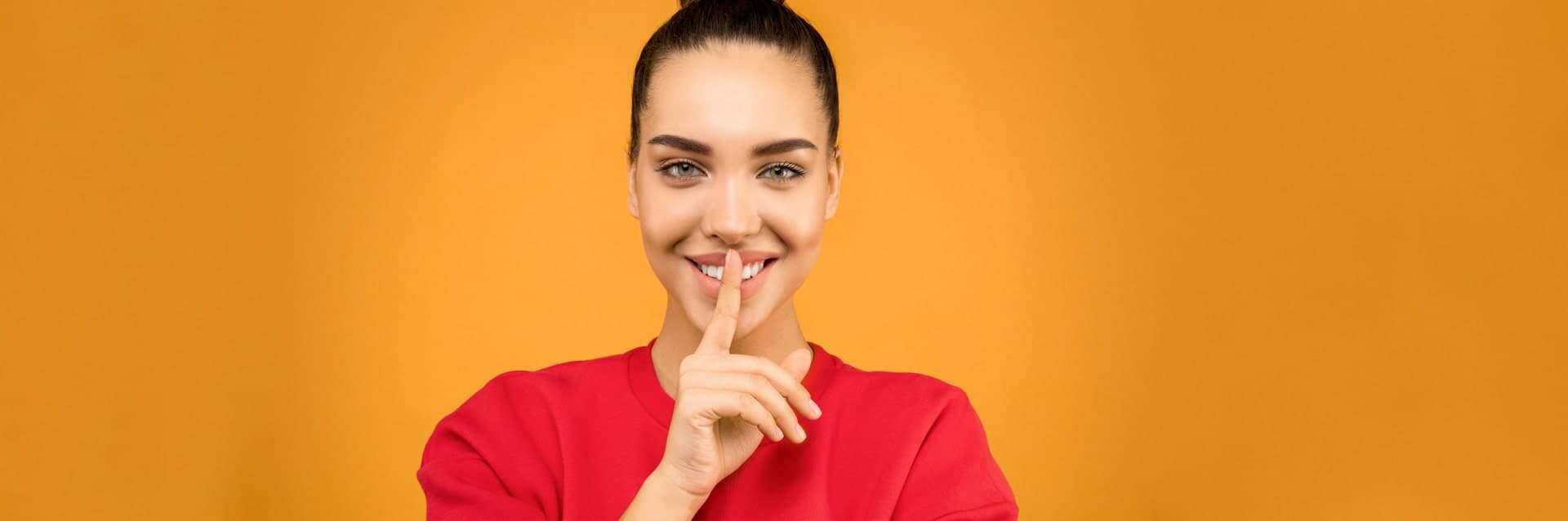 Smiling woman in a red shirt holding a finger to her lips in a â€œshhâ€ gesture, symbolizing confidentiality and discretion, representing a privacy policy concept.