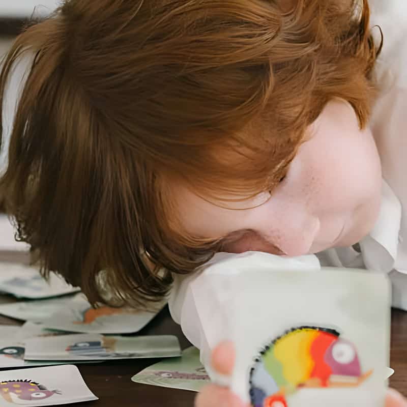 Child resting their head on a table while holding a colourful card, surrounded by playful illustrations â€” representing emotional exploration through play therapy.
