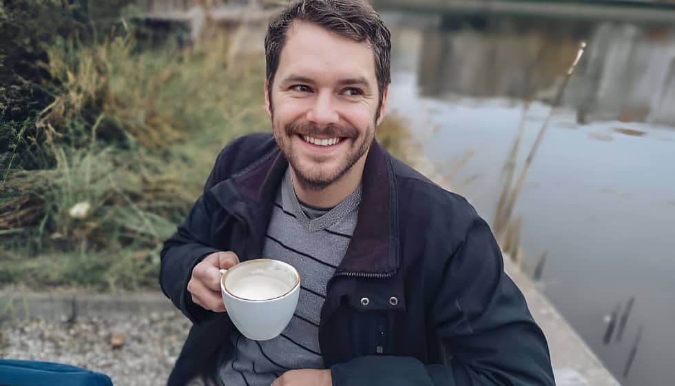 Smiling man holding a coffee cup by a peaceful mini-lake, representing the positive outcomes of working with the best EMDR therapist in Orangeville.