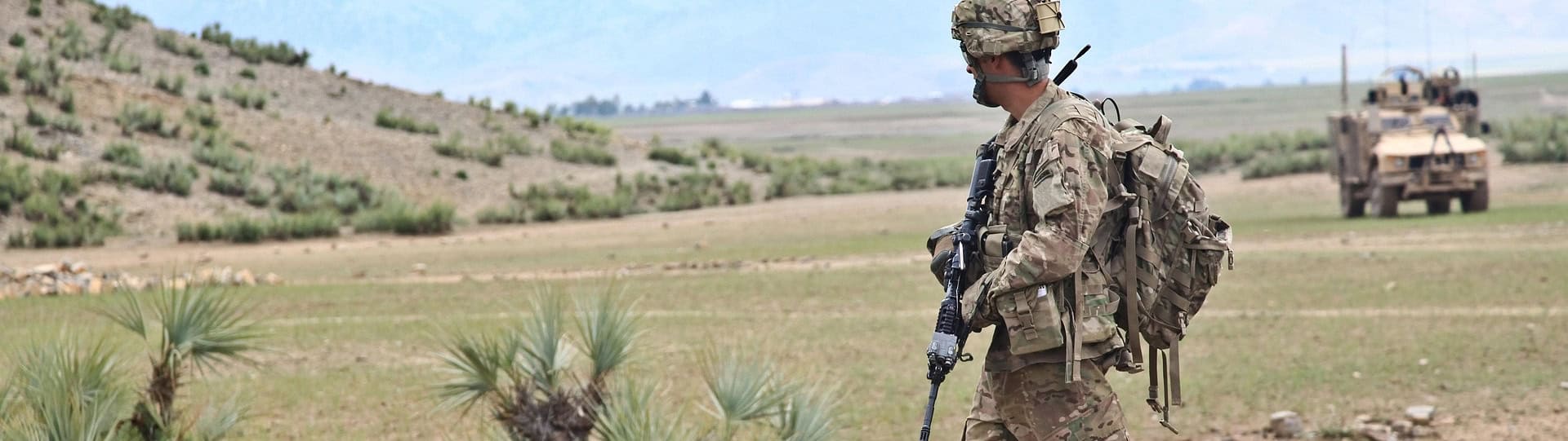 Soldier in full gear walking across a rugged landscape, in field, with military vehicles in the background, symbolizing the focus of EMDR Sessions for Military Veterans.