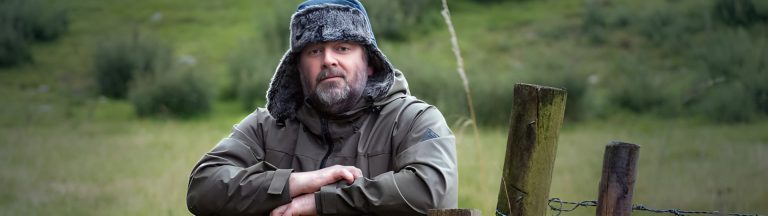 A man dressed for cold weather leans on a wooden fence in a rural setting, symbolizing personal reflection and lifestyle choices, ideal for illustrating in-person vs online EMDR therapy in Ontario.