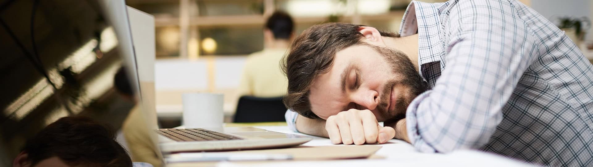 Man resting his head on his arms at a desk with eyes closed, practising mindfulness in a busy co-workspace to find calm and focus amidst daily stress.