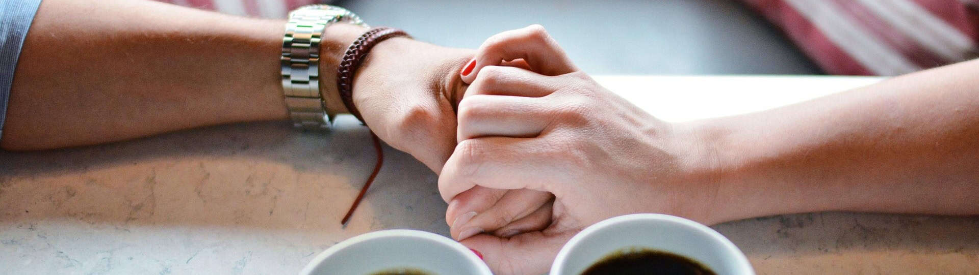Close-up of a couple holding hands across a table with coffee cups, symbolizing emotional connection and love maps in a relationship.